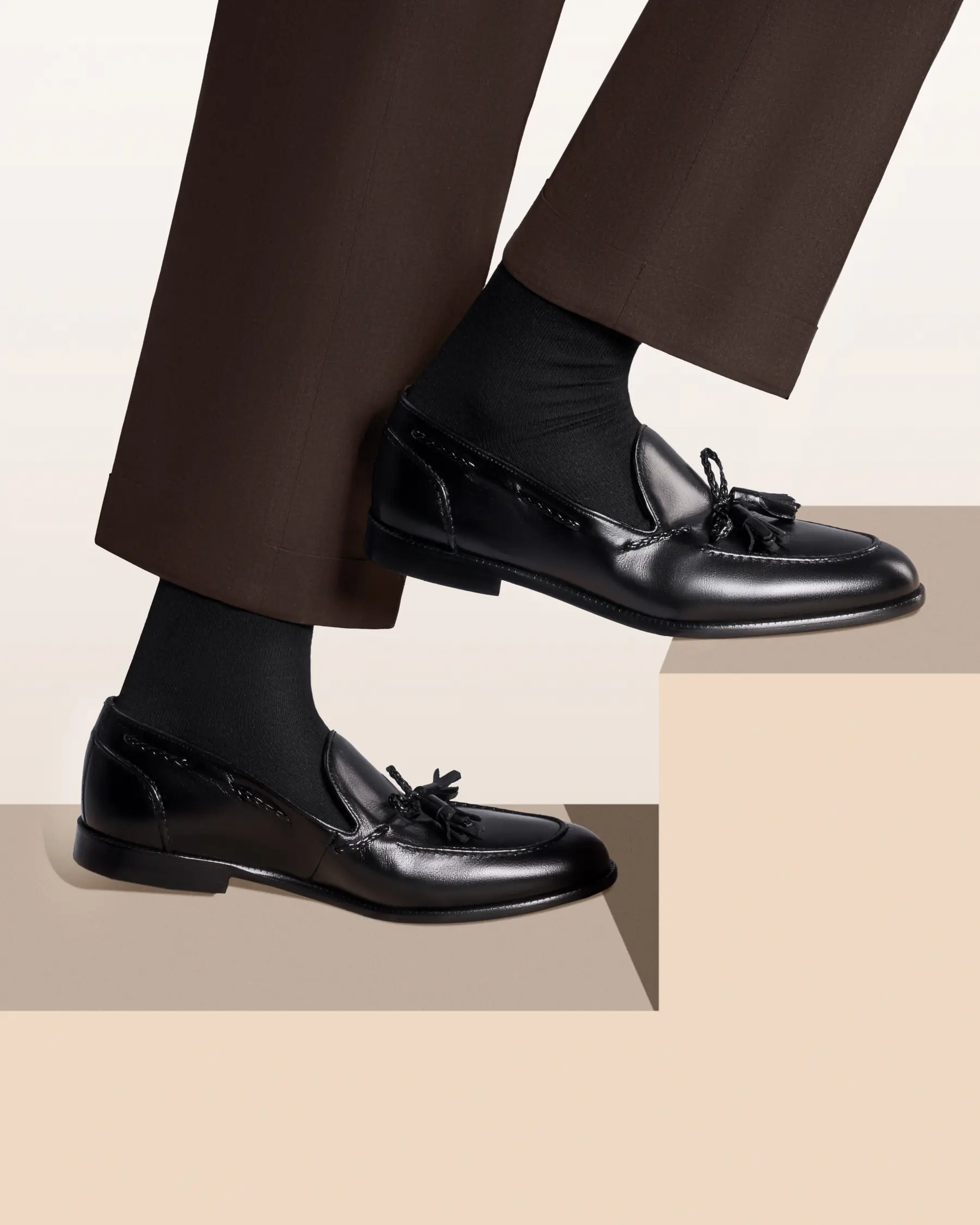 Black tassel loafers worn with brown trousers on cream steps